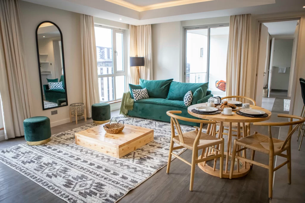 Teal sofa with patterned cushions, wooden coffee table with decor, round dining table with chairs, large patterned rug, and light-filled windows in a cozy living room setting