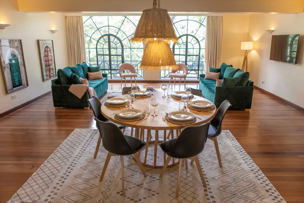 The dining room exudes modern elegance, with a wooden table elegantly set for dinner, surrounded by six sleek chairs featuring wooden legs and chic grey upholstery. Beneath the ensemble, a large woven area rug anchors the space, infusing it with warmth and texture. In the backdrop, inviting teal-colored sofas adorned with decorative cushions offer a cozy spot for relaxation.